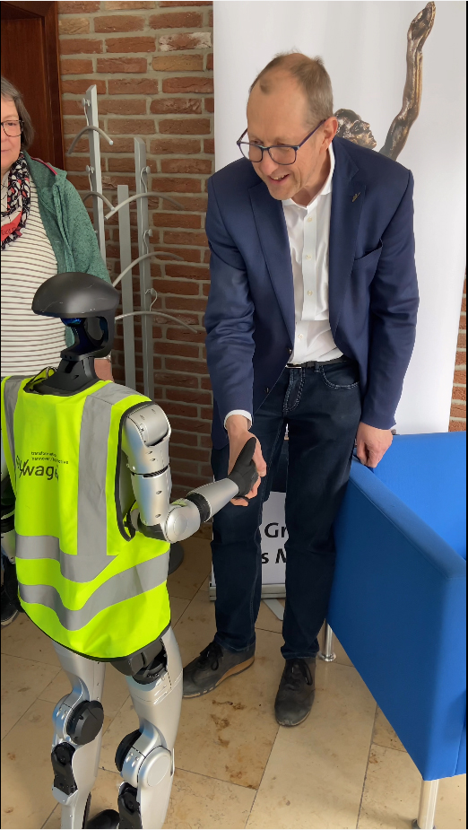 Robot in a safety vest shakes hands with a man in a suit, showcasing human-robot interaction in a professional setting.