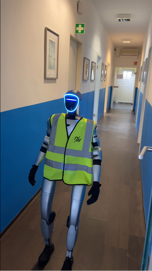 A humanoid robot in a reflective vest walking down a hallway with blue walls and framed artwork.