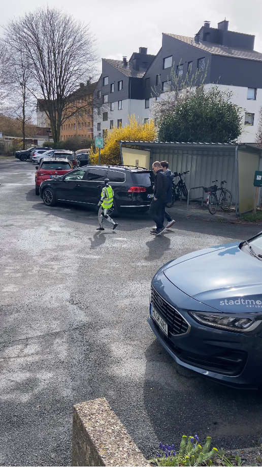 Two people walk in a parking area with cars and bicycles, while a child in a safety vest walks alongside them.