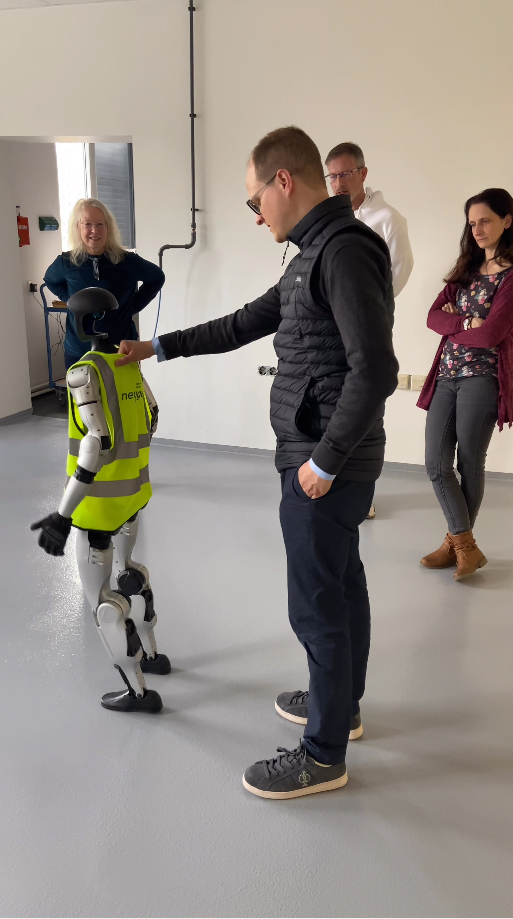 A man interacts with a humanoid robot in a demonstration, while onlookers observe in a bright indoor space.
