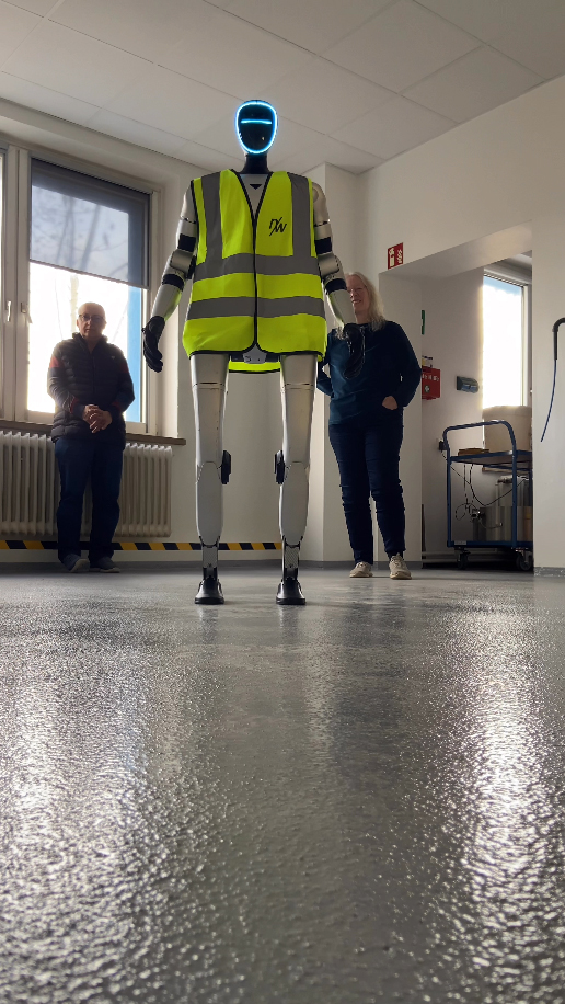 A humanoid robot in a safety vest stands in a room with onlookers, showcasing advanced technology and innovation.