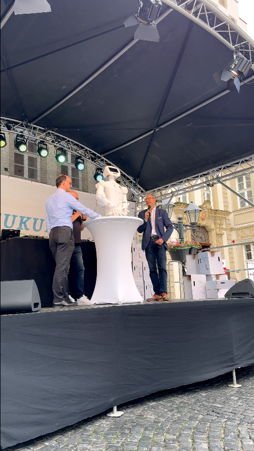 Men on stage presenting a robot during an outdoor event, featuring a festive atmosphere and bright lighting.