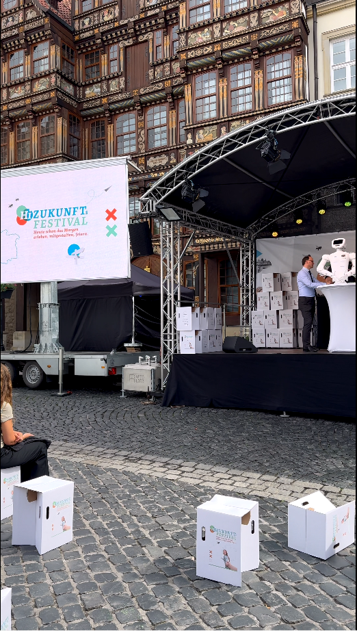 Zukunft Festival stage featuring speaker, logo display, and audience seating in a vibrant outdoor setting.