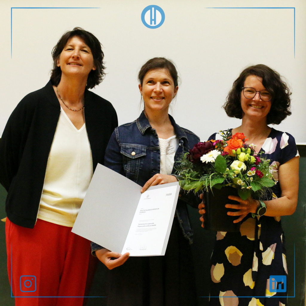 Three women celebrate a milestone, holding a certificate and a bouquet of flowers at a formal event.